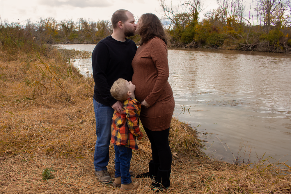 Family maternity moment by riverside with autumn foliage in background.