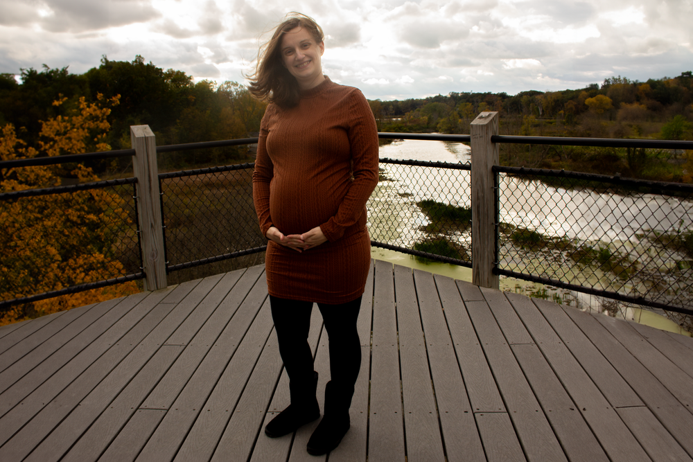 A person in a burgundy sweater stands on a wooden deck overlooking an autumn lake with fall foliage.
