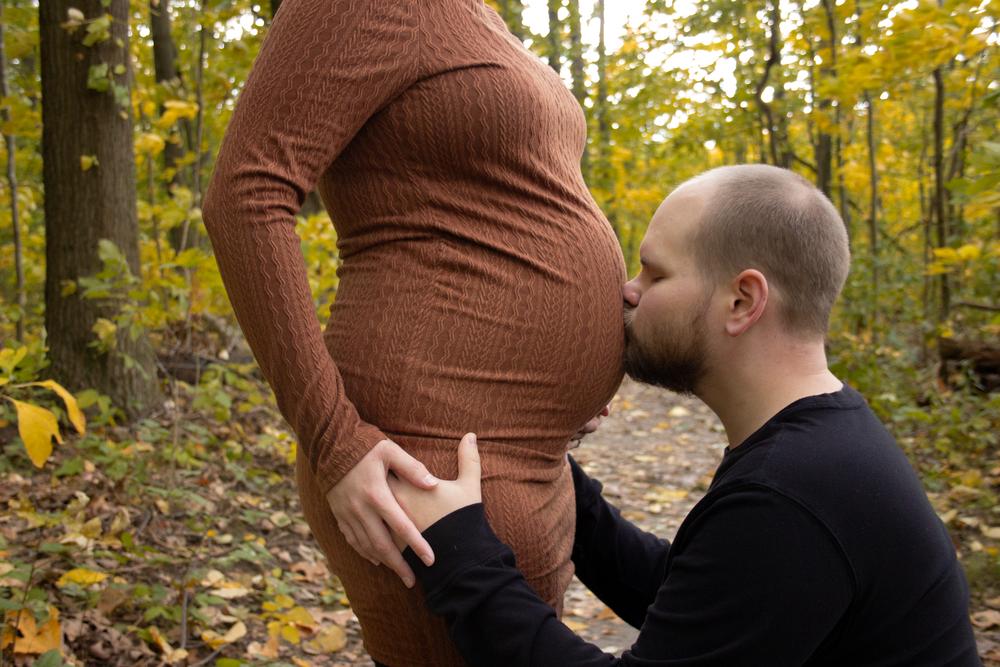 A tender maternity moment captured in autumn woods with fallen leaves and warm lighting.