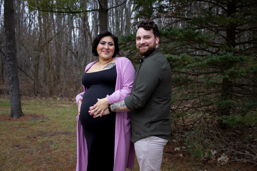 A couple shares an intimate maternity moment among evergreen trees in a forest setting.