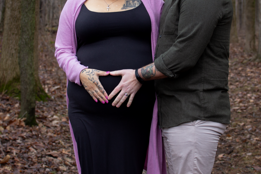 A maternity photo showing hands forming a heart shape on a pregnant belly in a forest setting.