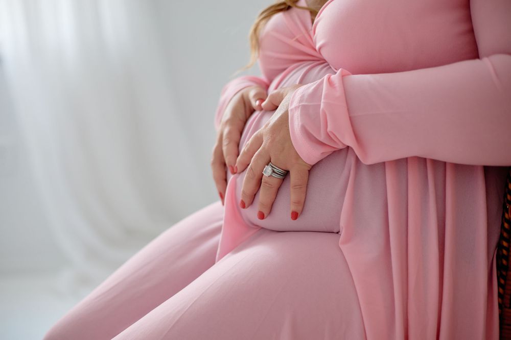 Gentle hands rest on pregnant belly wearing soft pink maternity dress.