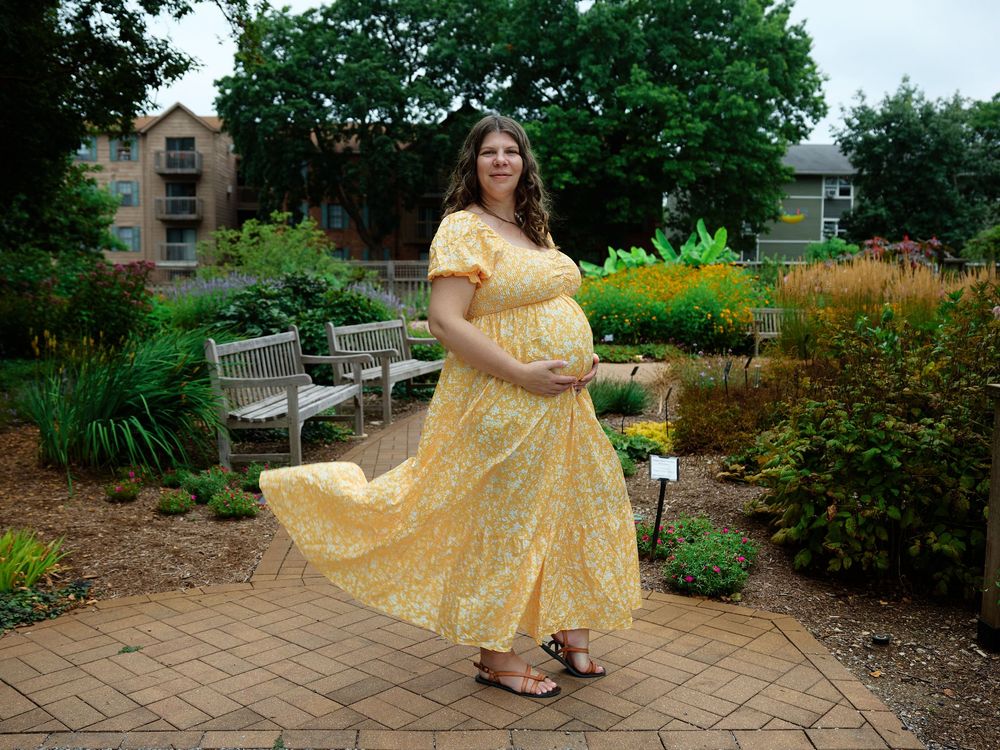 Pregnant woman in yellow floral maxi dress posing in a garden park with benches and greenery.