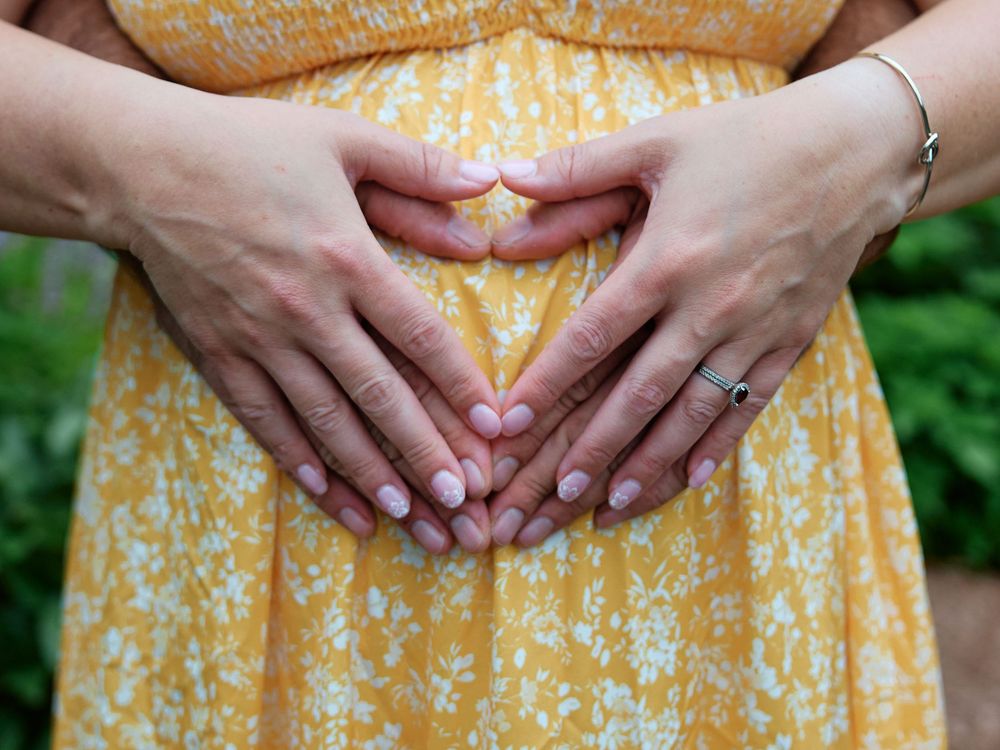 Pregnant woman in yellow floral dress with hands forming heart shape on baby bump.