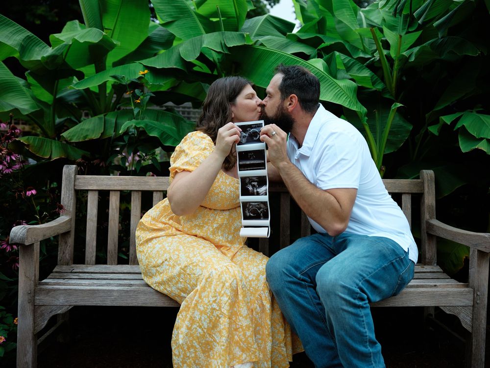 Couple kissing on garden bench holding ultrasound photos, pregnancy announcement in lush green tropical setting.
