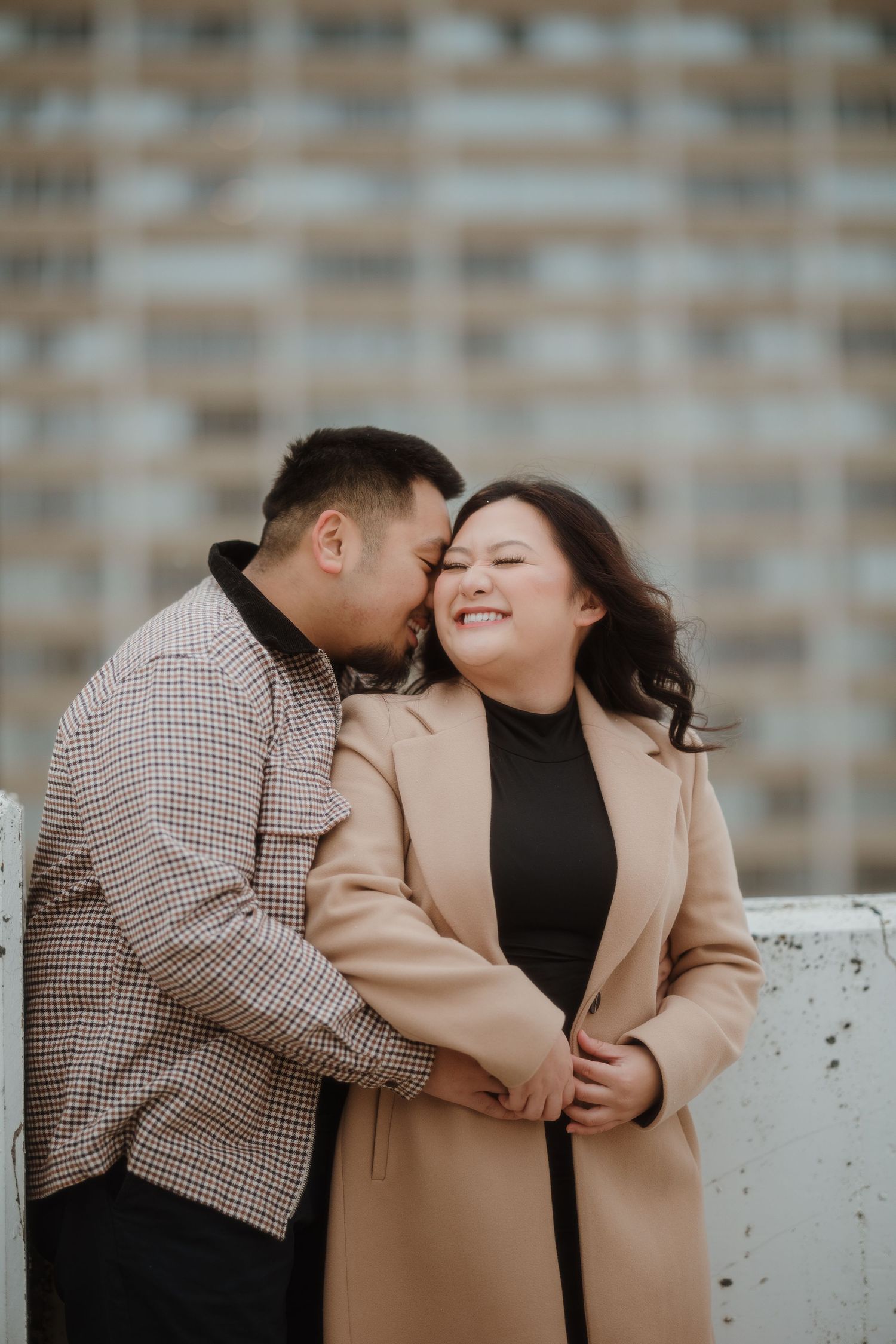 A romantic couple shares an intimate moment outdoors, with the woman wearing a camel coat and black top against an urban backdrop.