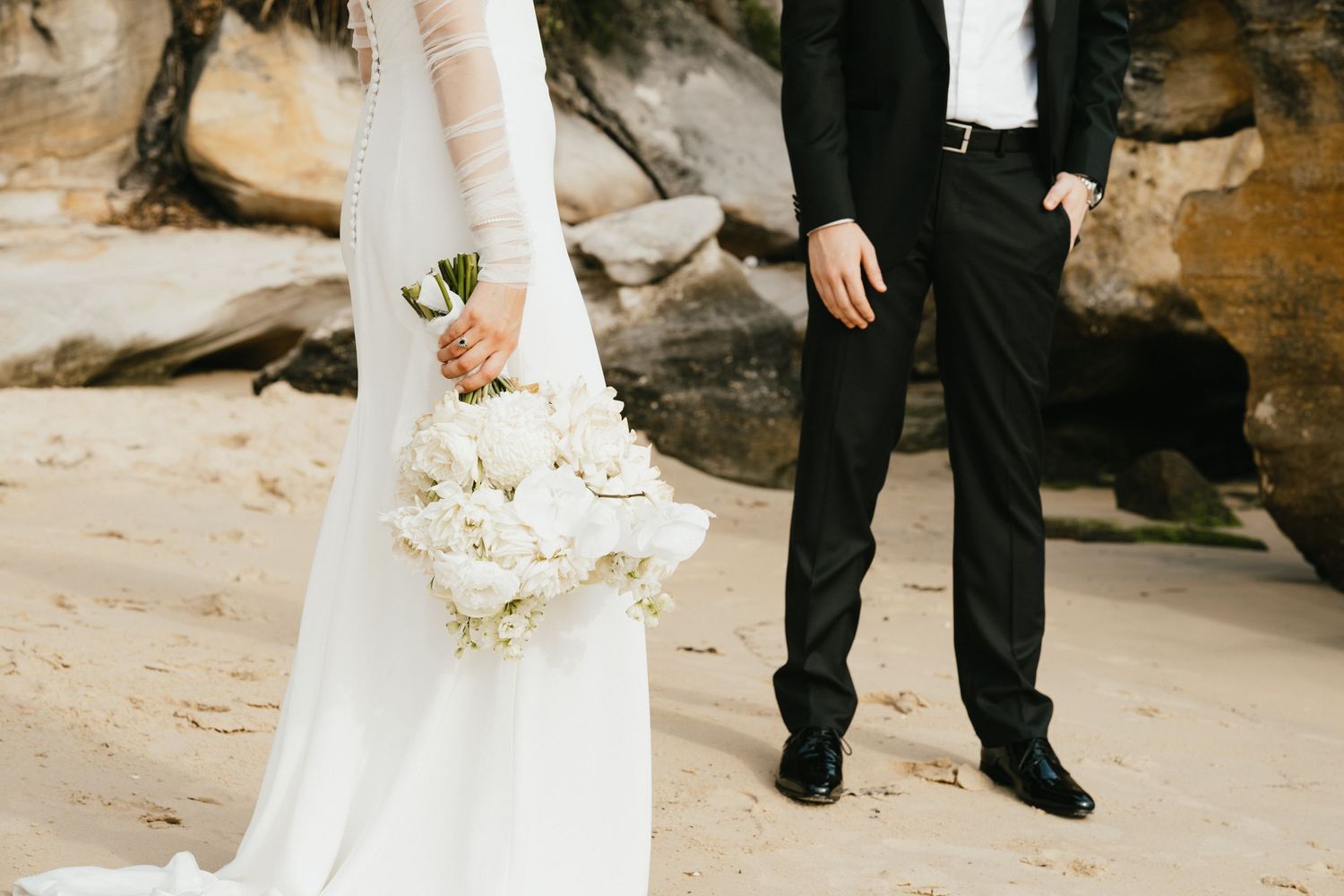 A wedding couple standing on a beach with the bride holding a white floral bouquet against rocky cliffs.