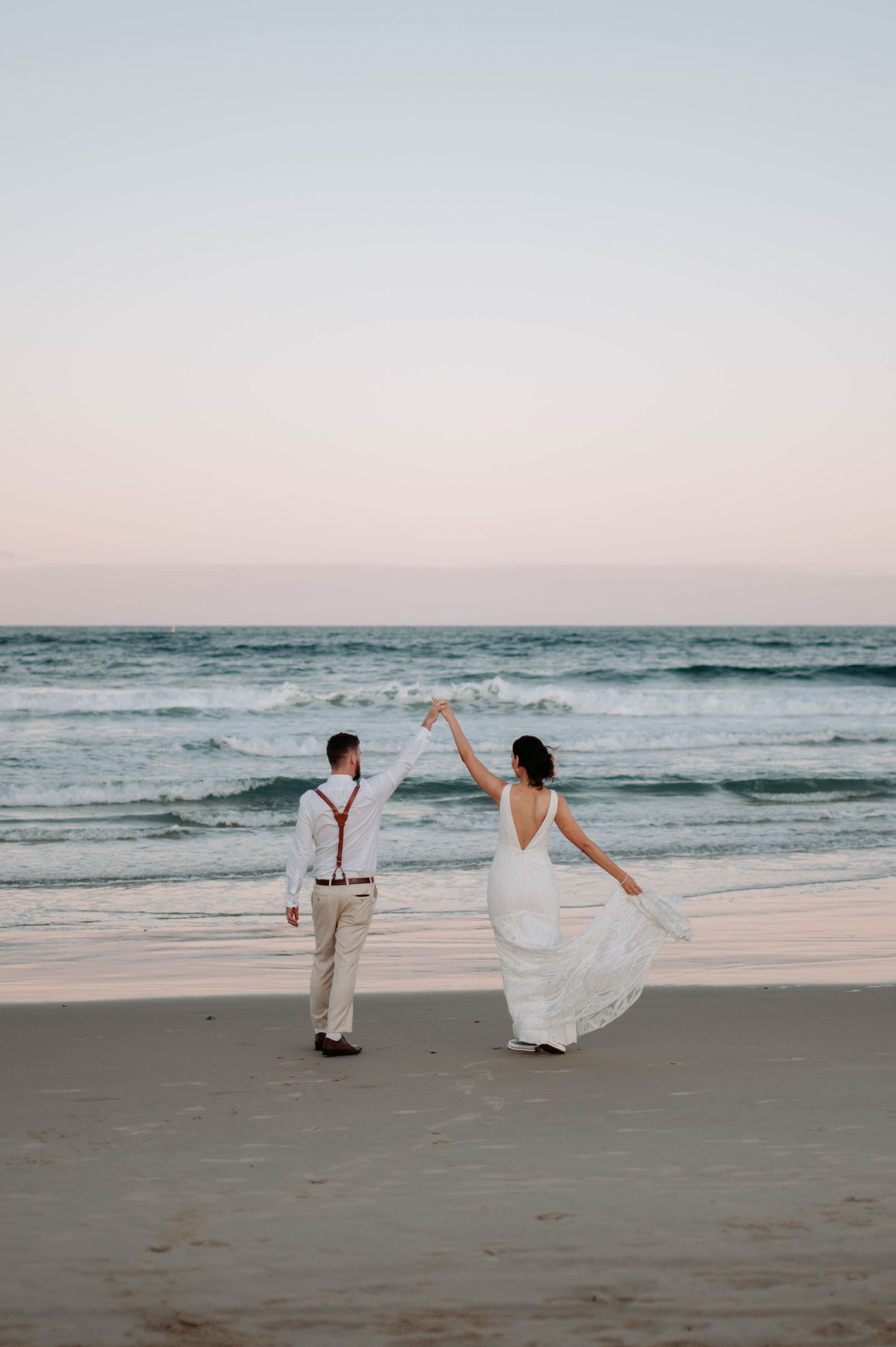 Couple dancing and twirling on beach at sunset in wedding attire.