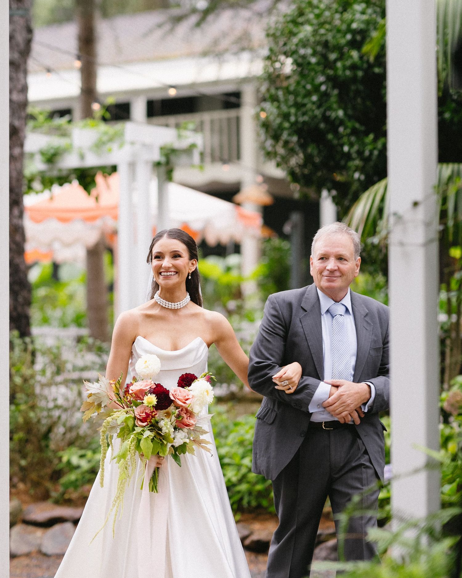 Bride in strapless gown walking arm-in-arm with father figure down garden path.