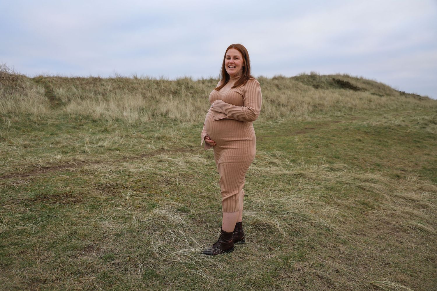Expectant mother in a beige ribbed dress stands on grassy hillside during pregnancy photoshoot.