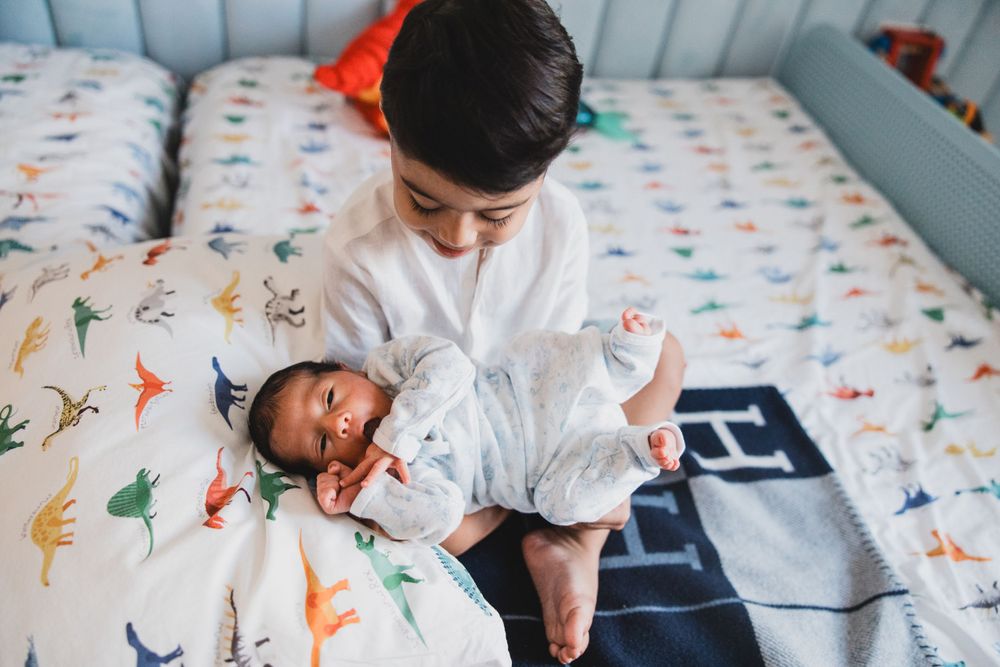 Two children share a playful moment on a colorful dinosaur-patterned bedsheet.