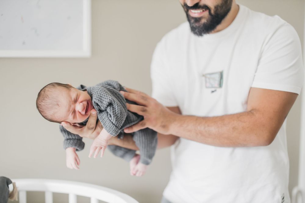 Caregiver gently holds sleeping newborn wearing grey onesie near white crib.