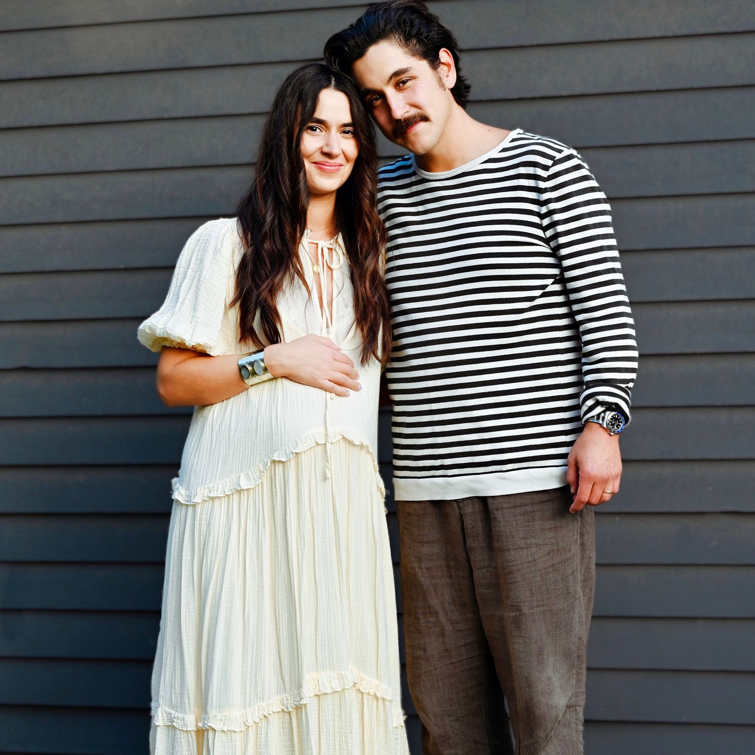 Couple in casual wear standing against grey wall outdoors.