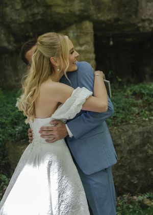 Newlyweds share an intimate embrace outdoors, with bride in white gown and groom in blue suit.