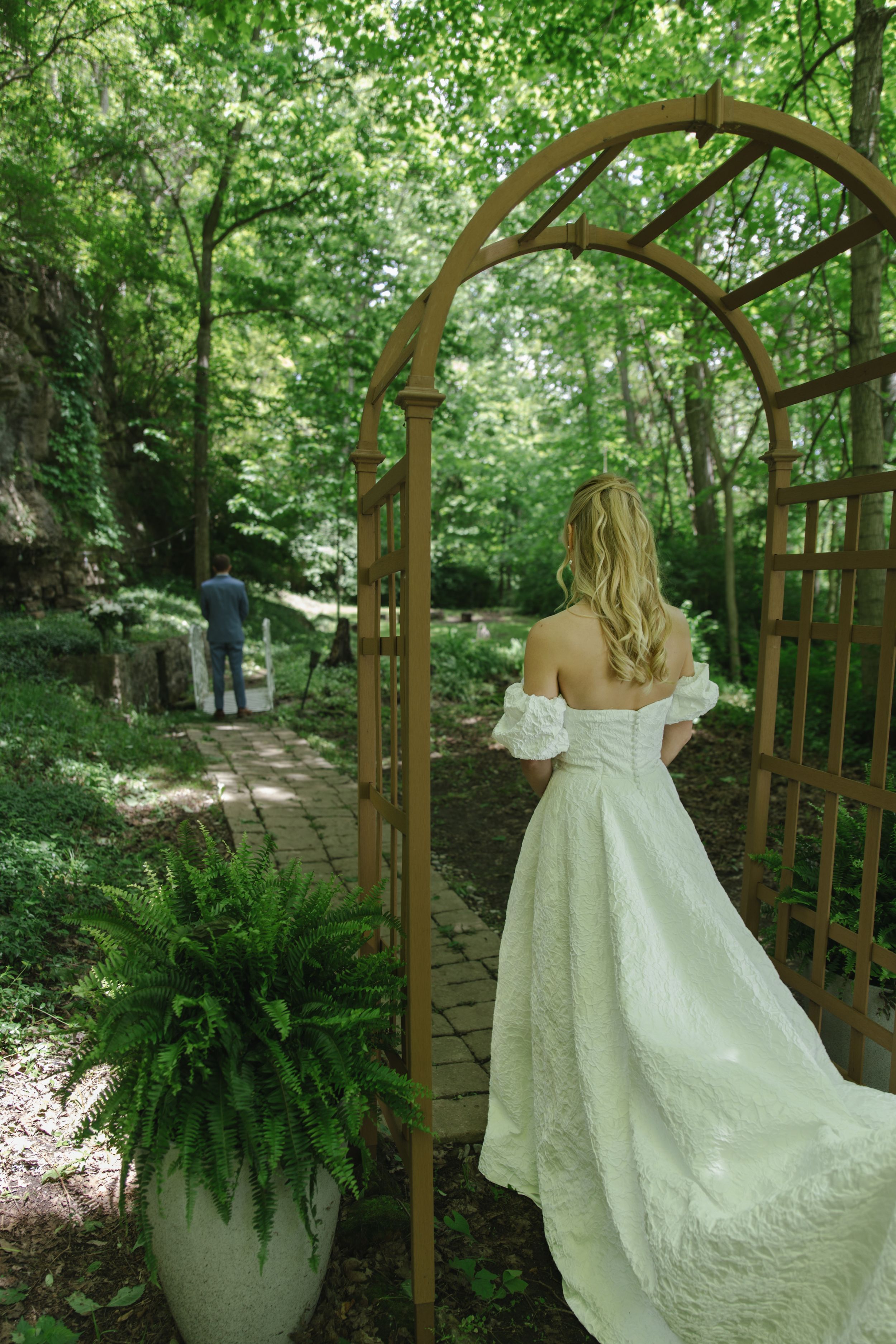 A bride in an off-shoulder wedding gown walks through a garden arch pathway.