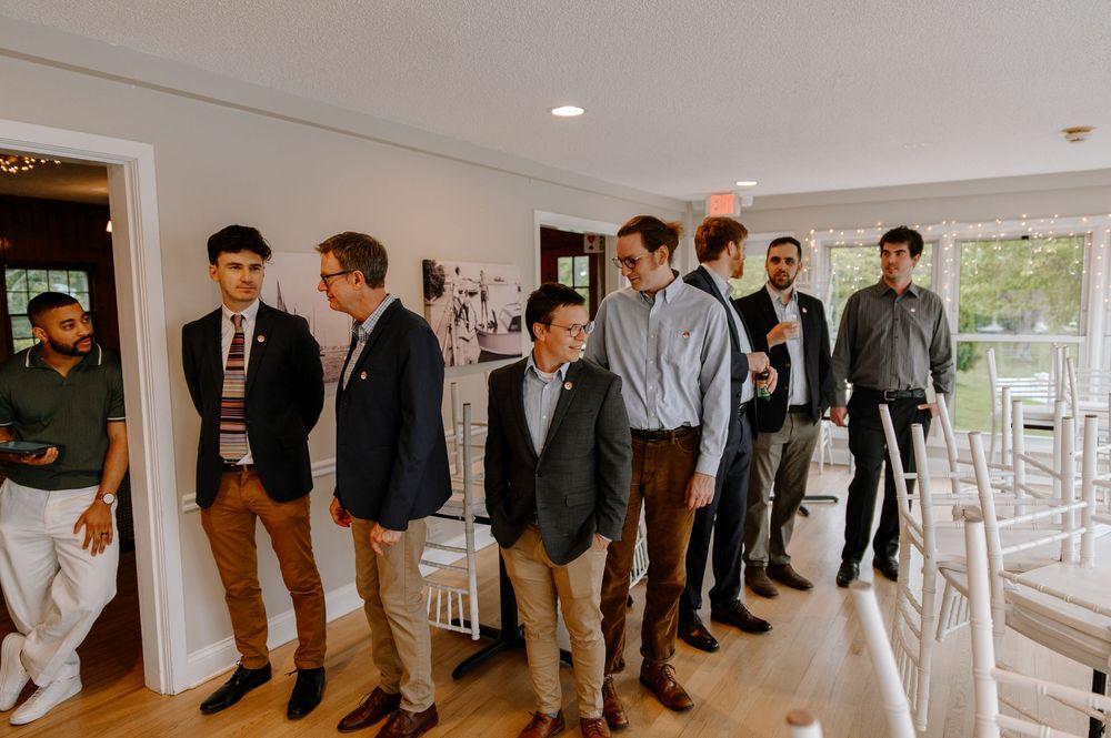 A group of men in casual and business casual attire standing in line against a white wall in a bright interior space.