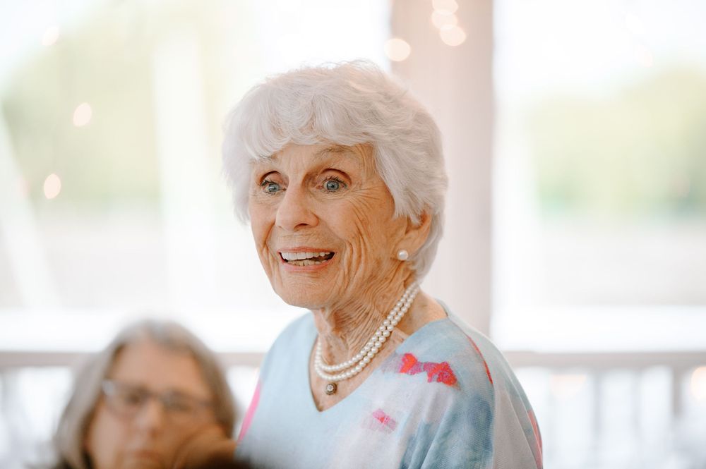 An elderly woman in a light blue sweater with pink accents sits on a porch smiling warmly in natural daylight.