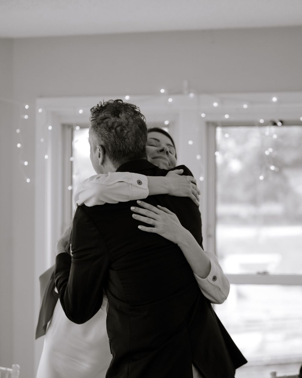 Two people share an intimate embrace under twinkling string lights in a black and white photograph.