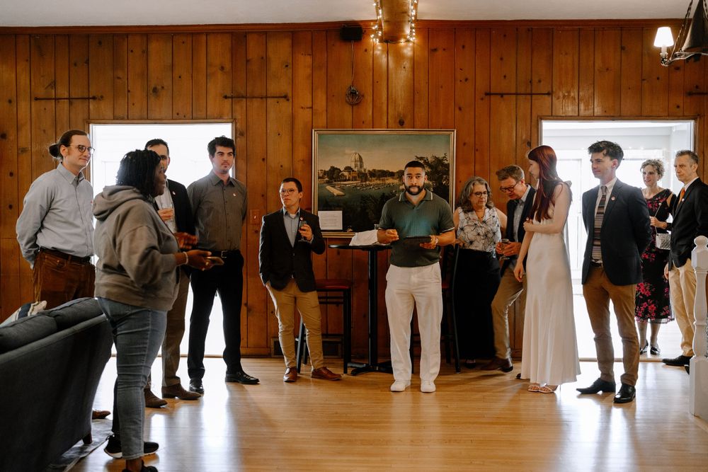 A group of people gather around a piano in a wood-paneled room during what appears to be a social event.