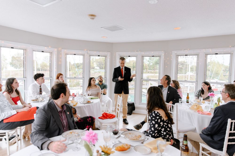 A group of wedding guests sit at round tables in a bright reception room while a person stands making a toast.