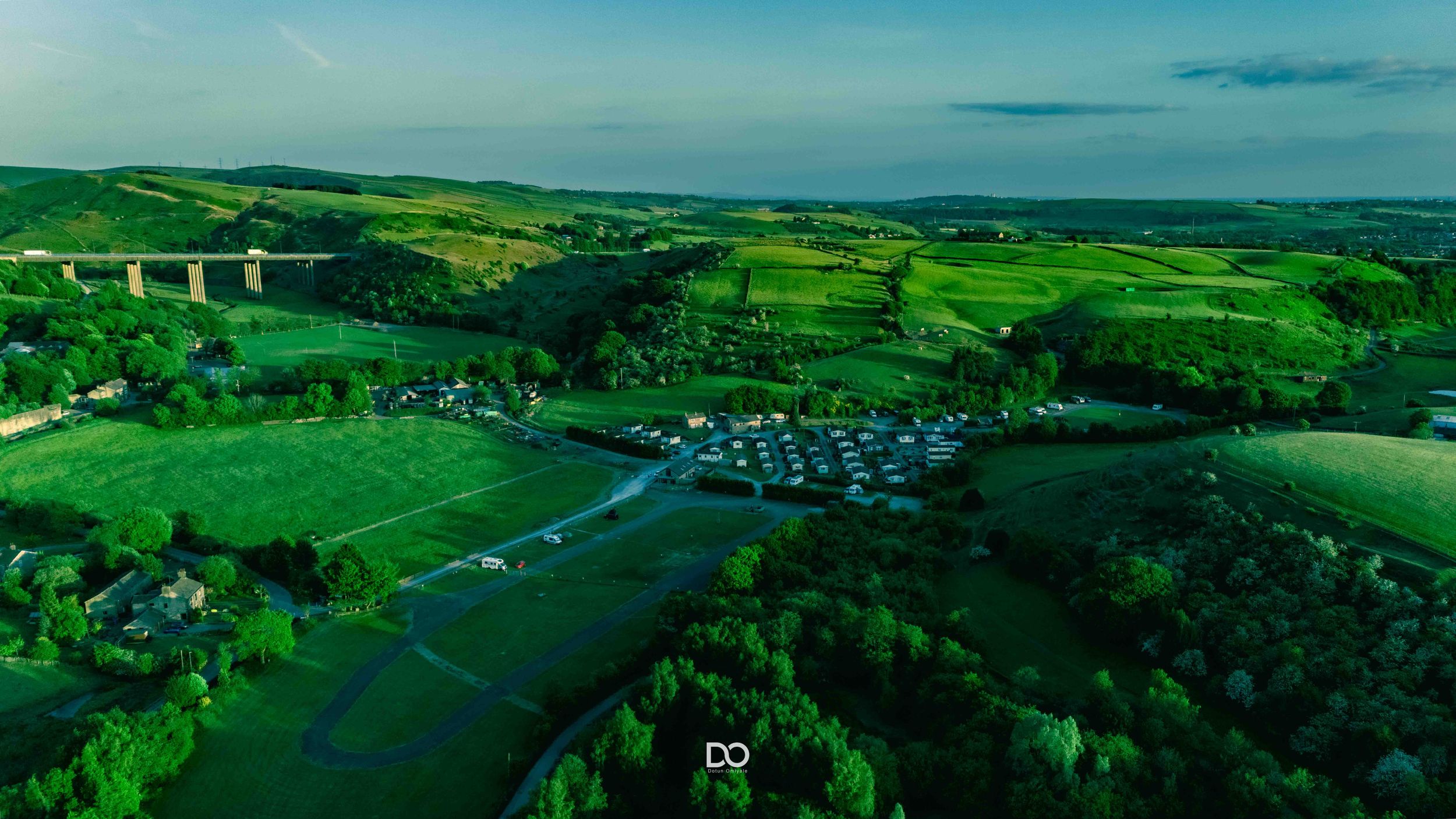 Aerial panorama of lush green countryside with rolling hills, farm fields and scattered buildings stretching to the horizon.