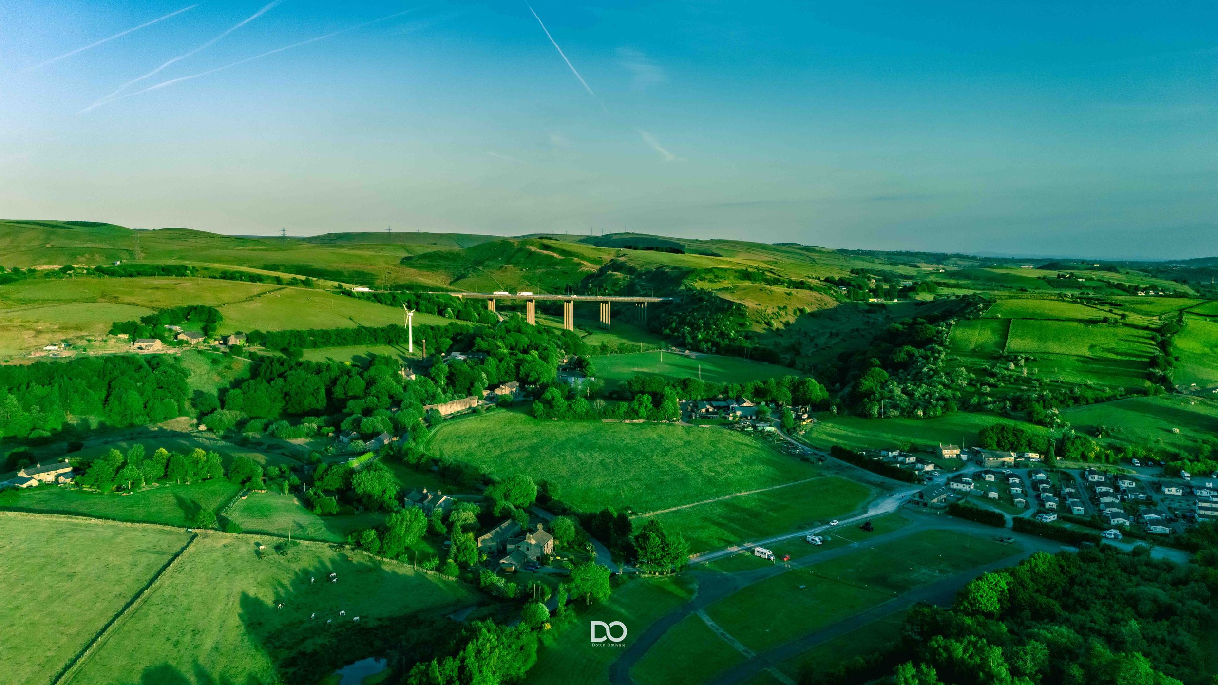 Aerial view of green countryside with rolling hills, farmland, and a tall stone viaduct bridge spanning a valley.