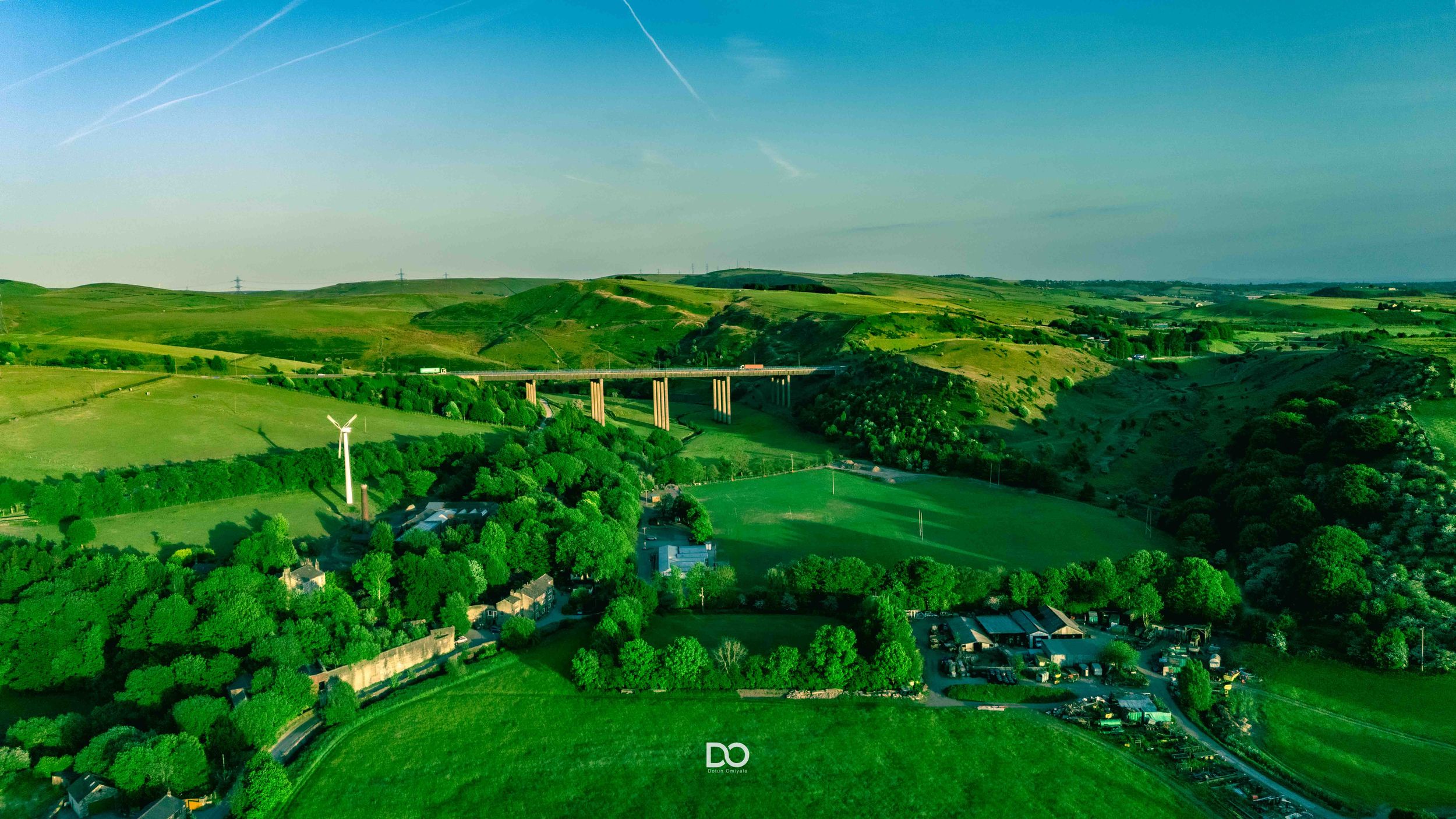 A scenic aerial view of a rural village nestled in rolling green hills with a railway viaduct crossing the valley.