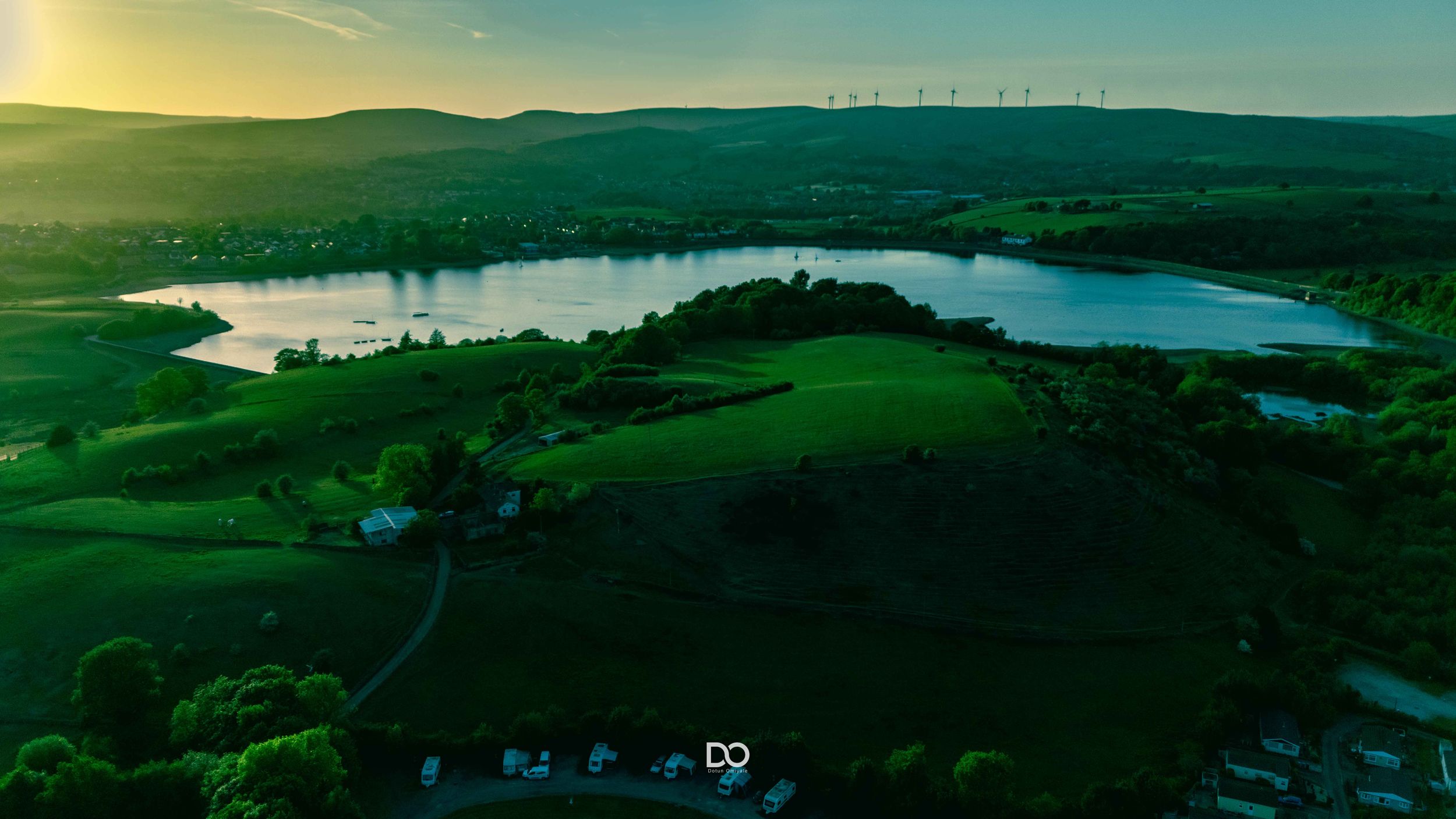 Dusk aerial shot of a calm lake nestled in green hills with distant wind turbines.