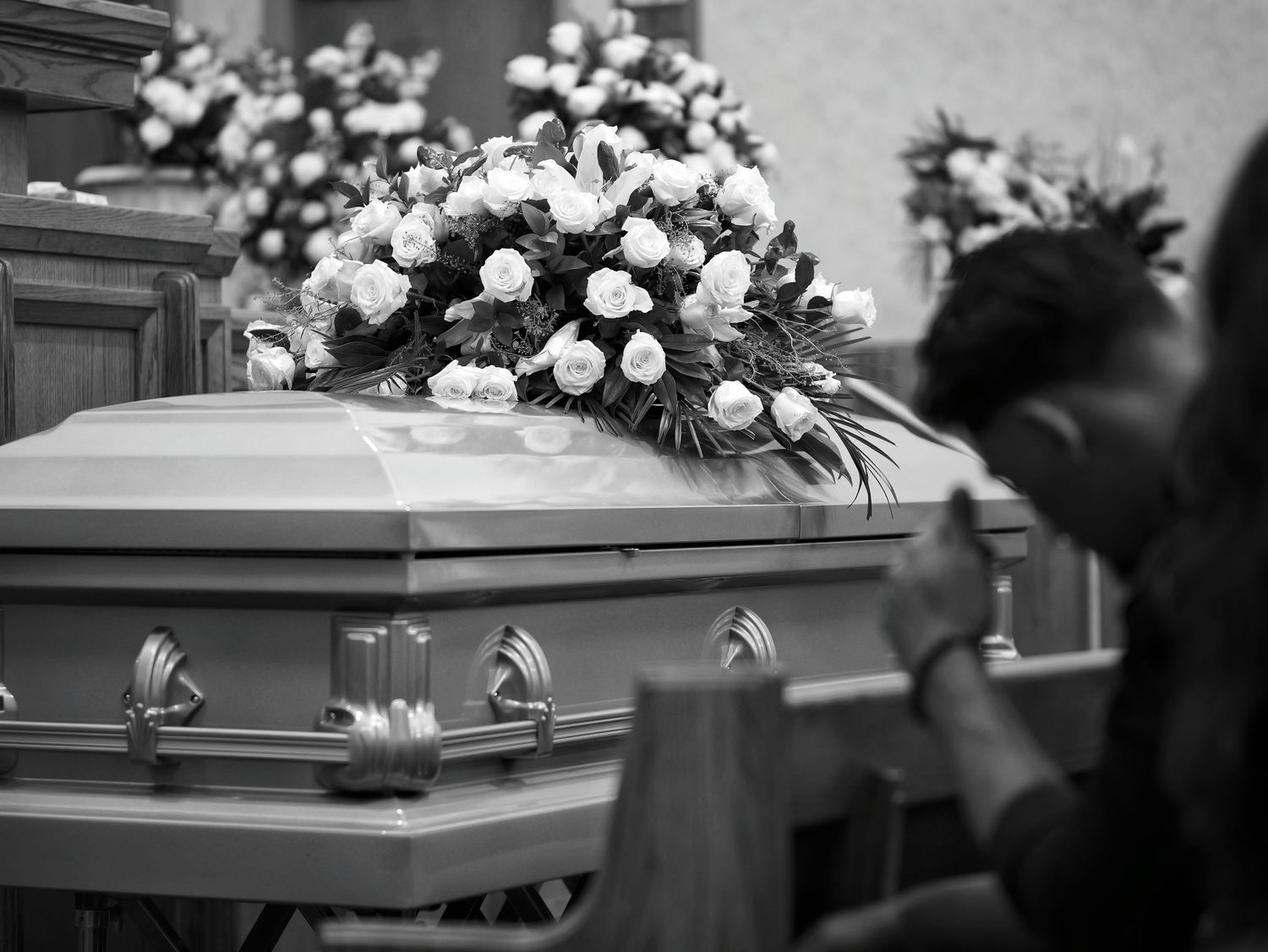 a beautiful arrangement of flowers sit atop the casket in a chapel. The son of the woman holds his head in his hands.