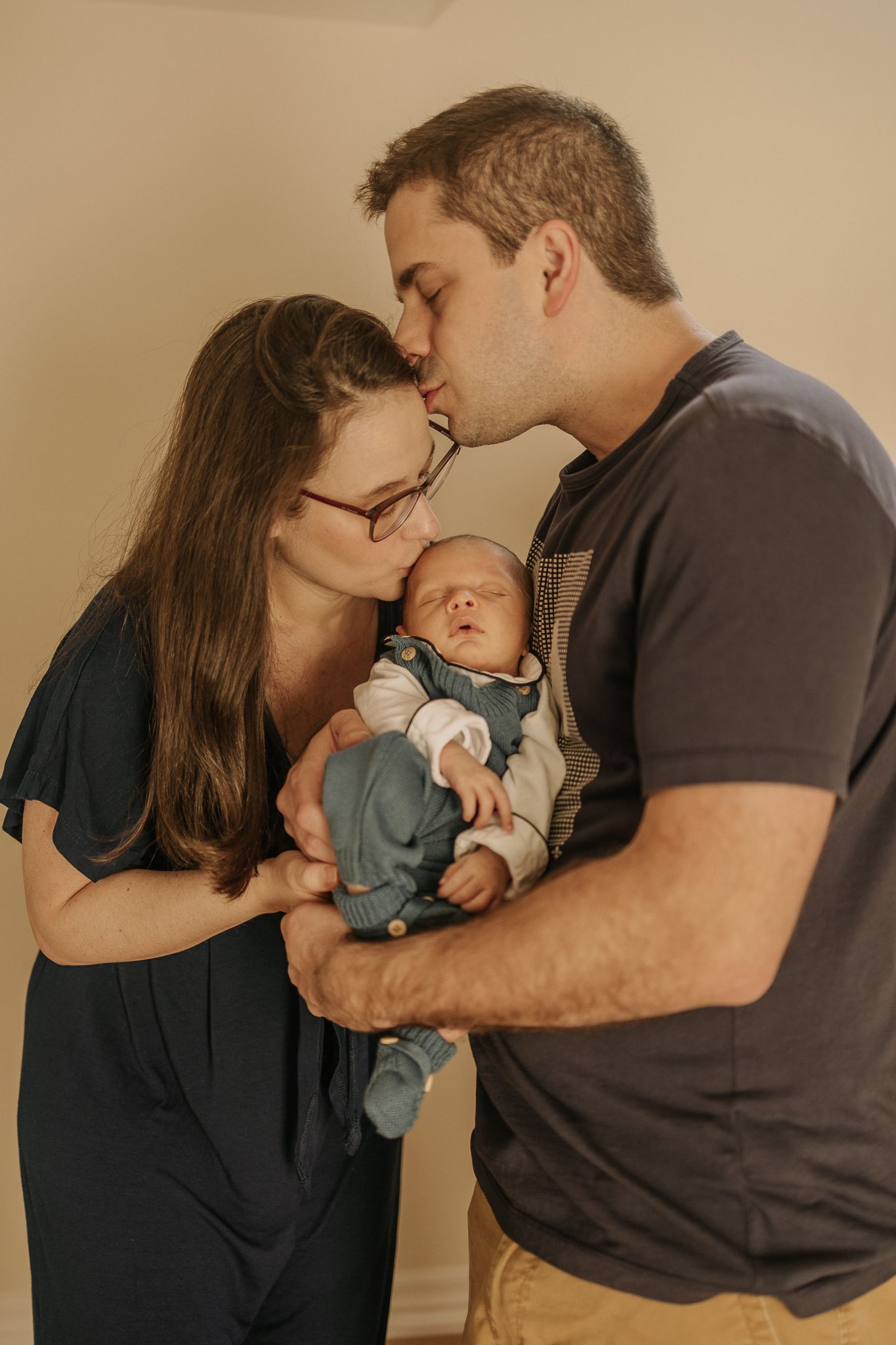 Parents holding their newborn baby, where. the mom is kissing the baby's head and the father is kissing the mom's head