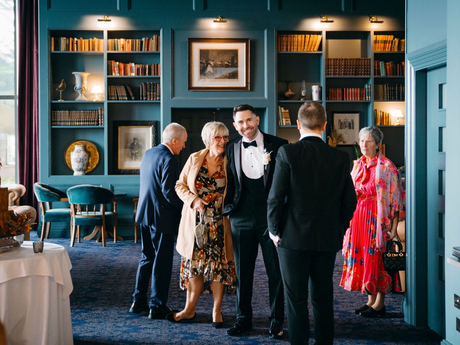 Panoramic view of well-dressed guests mingling in a dimly lit library space during an elegant evening event.