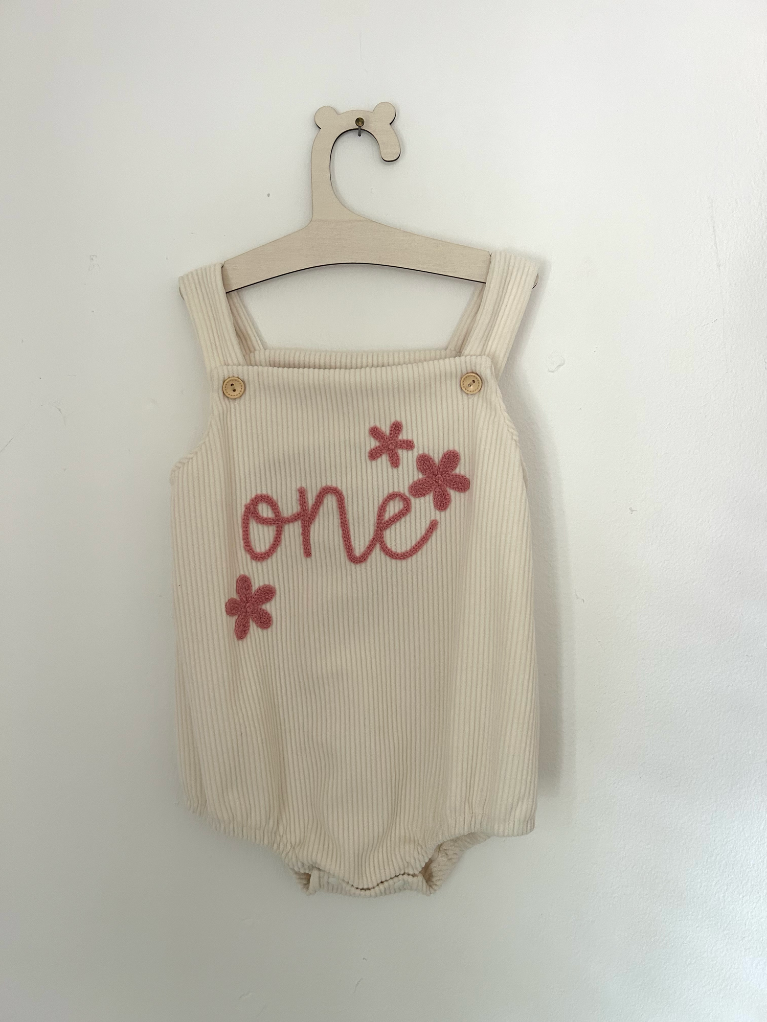 White knit baby romper with 'ONE' text and small bow detail in pink embroidery against hanging clothing background.