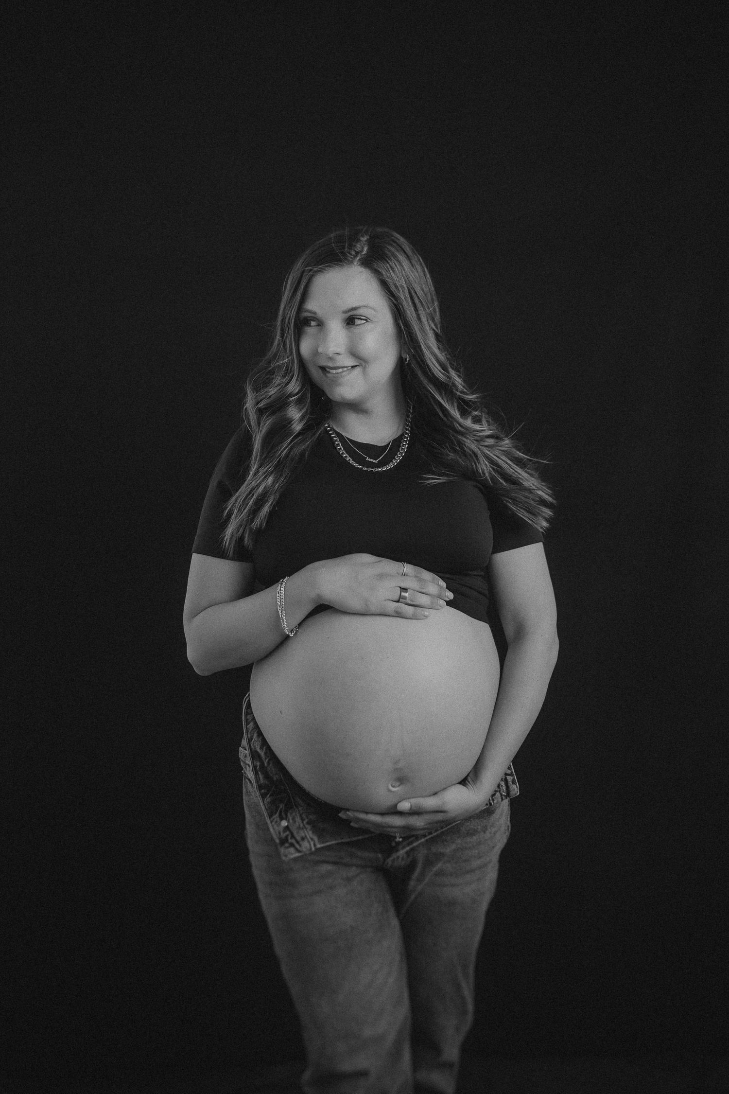 Black and white maternity portrait of an expectant mother in jeans and t-shirt against dark background.
