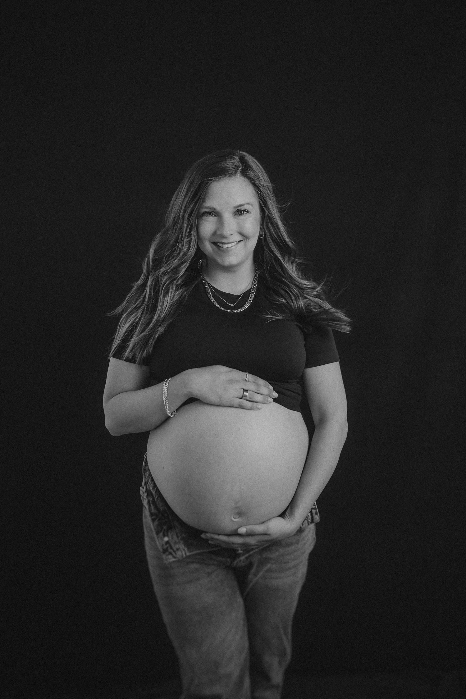 Black and white maternity portrait shows an expectant mother in casual attire against a dark background.