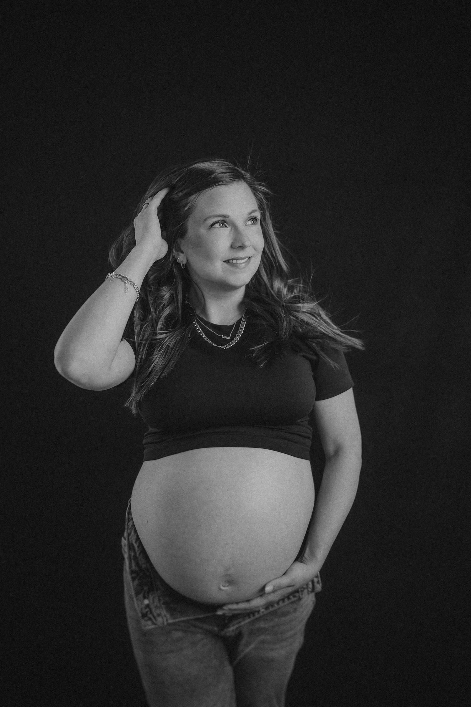 Black and white maternity portrait of an expectant mother in a dark shirt and jeans against a dark background.