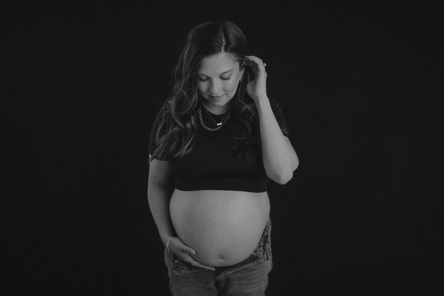 Black and white photo showing an expectant mother in dark clothing touching her bare pregnant belly against dark backdrop.