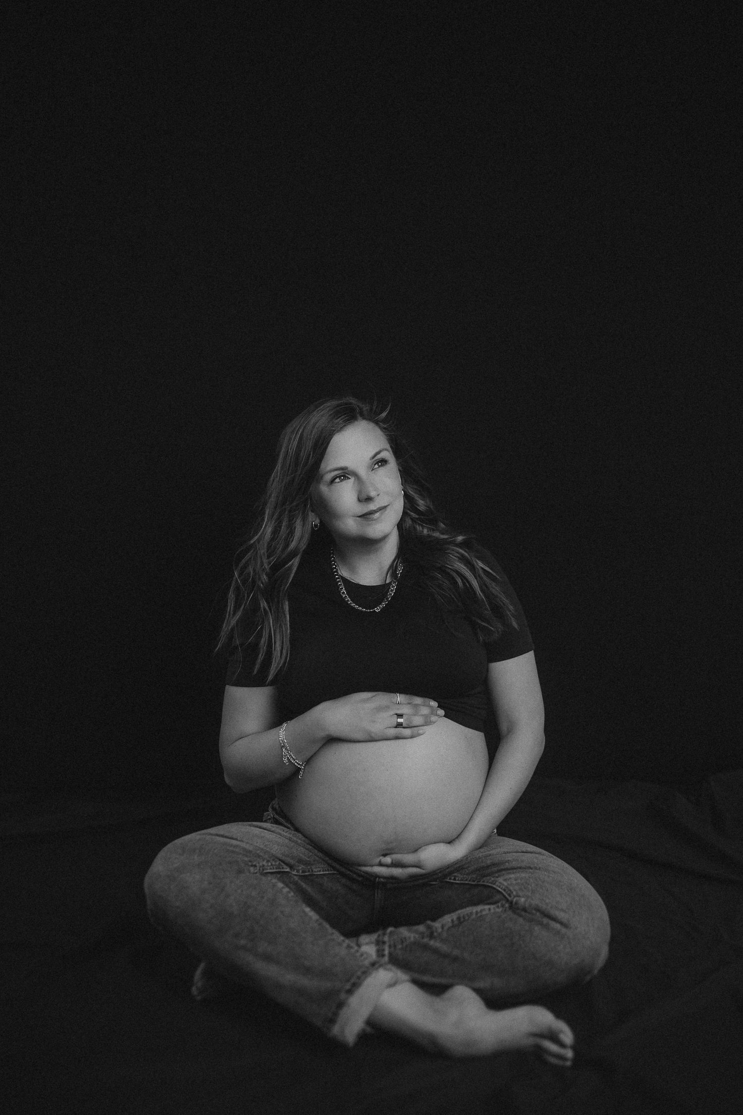 A black and white maternity portrait shows an expectant mother sitting cross-legged in a dark studio setting.