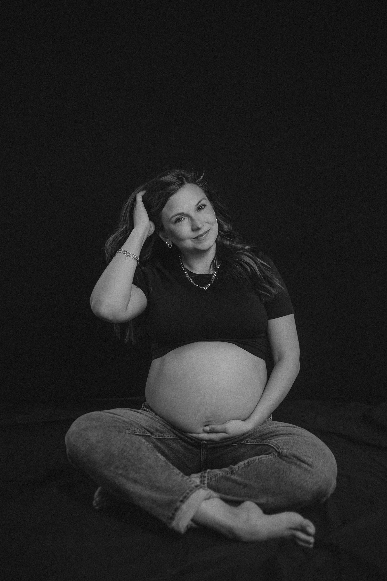 Artistic black and white maternity portrait of an expectant mother sitting cross-legged in casual attire against dark background.