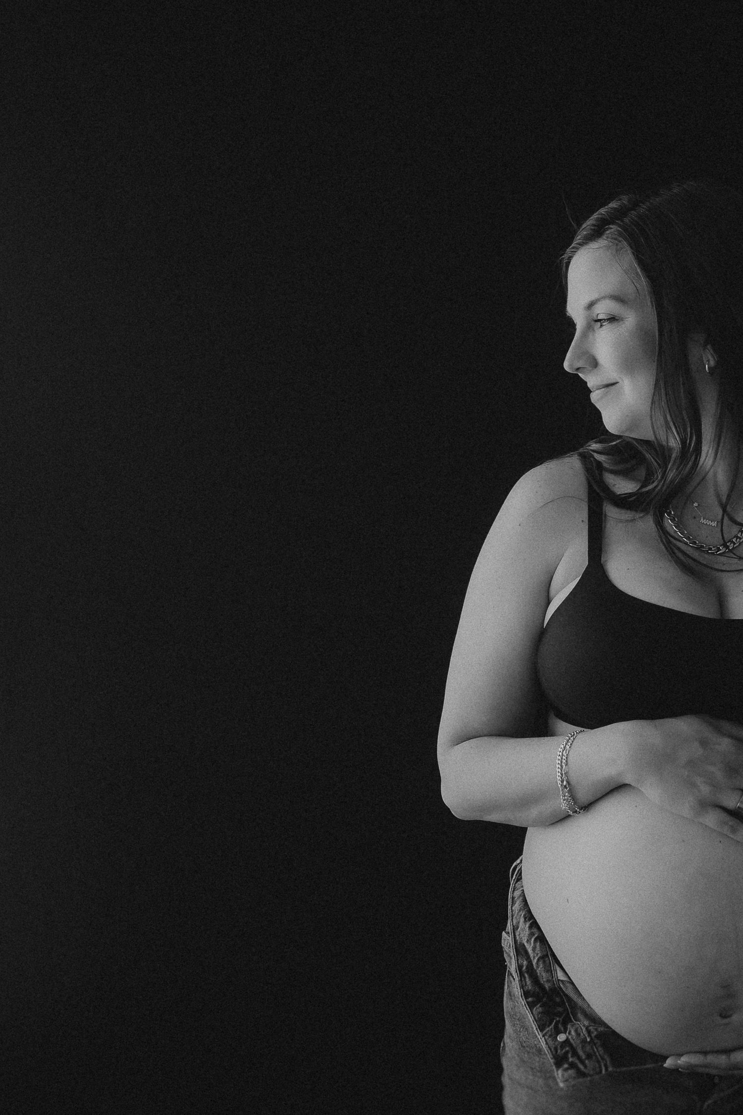 Black and white maternity portrait showing side profile of pregnant woman in dark top against dramatic black background.
