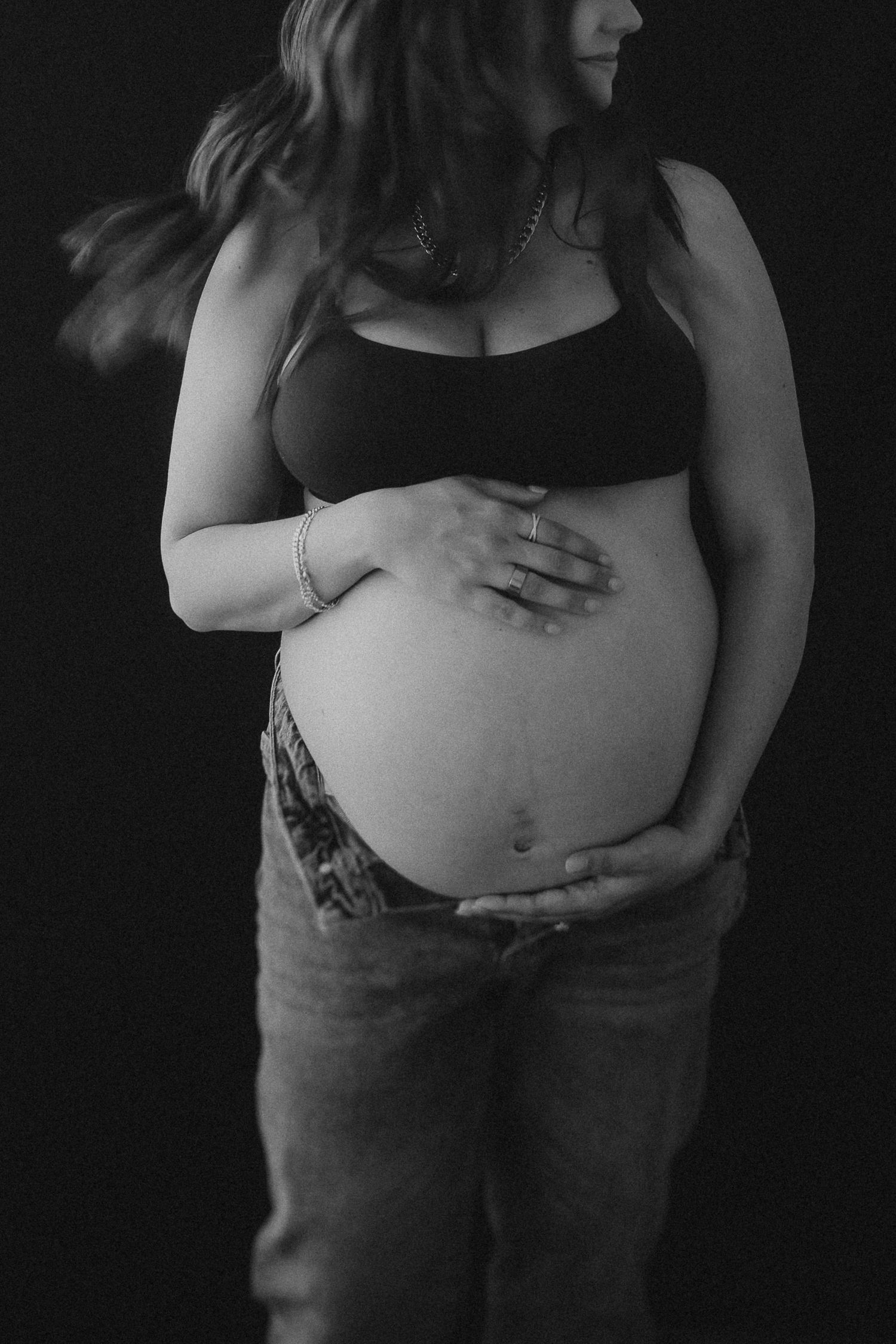 Black and white maternity photo of pregnant person in crop top and jeans cradling their belly against dark background.