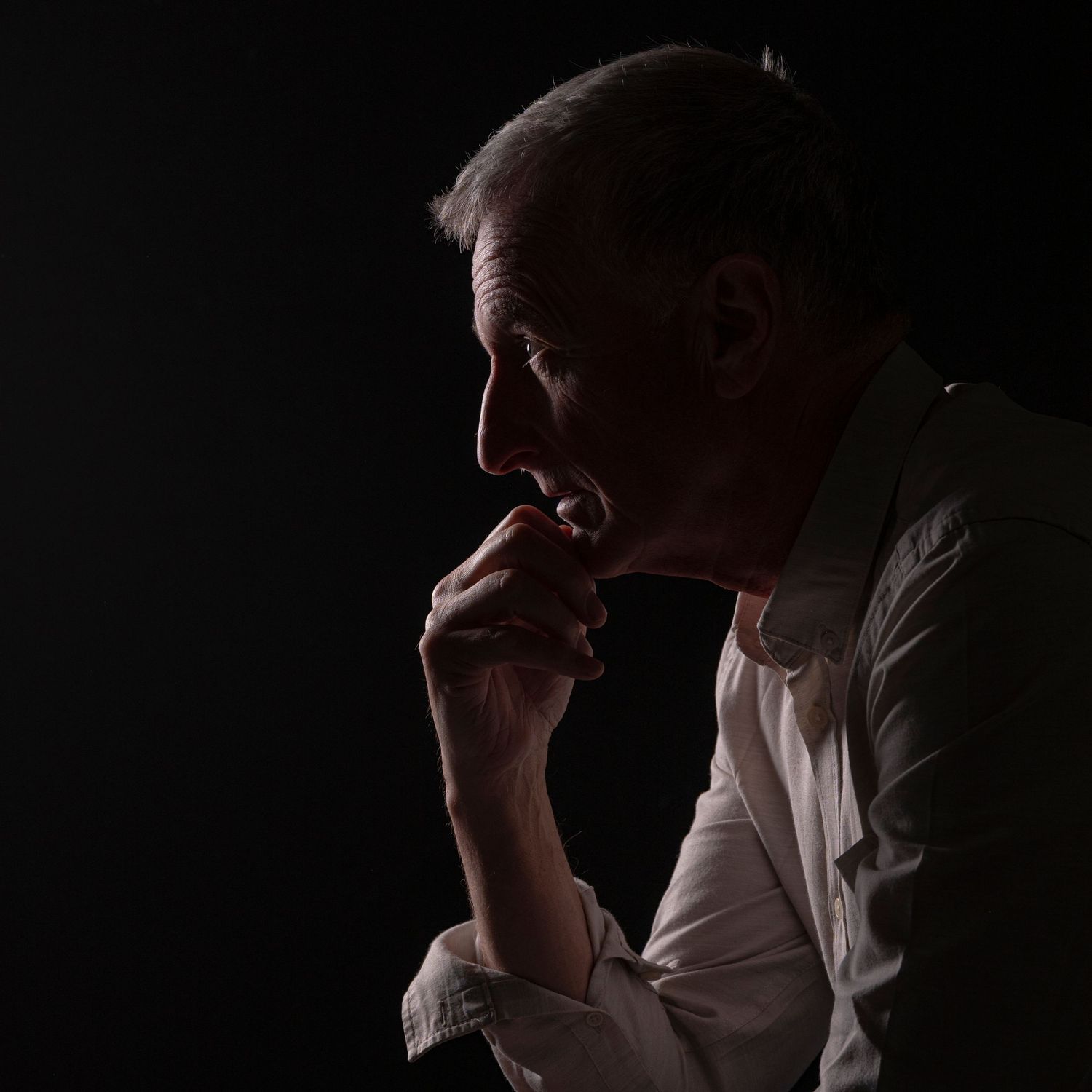 Elderly man in contemplative pose against dark background.