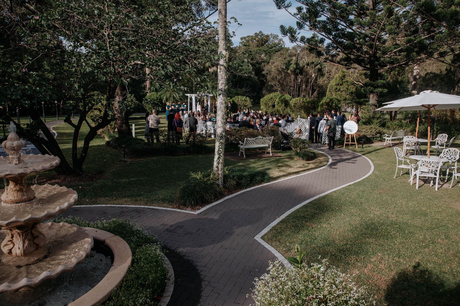 A scenic outdoor wedding ceremony setup with white chairs arranged on a grassy lawn surrounded by tall trees.