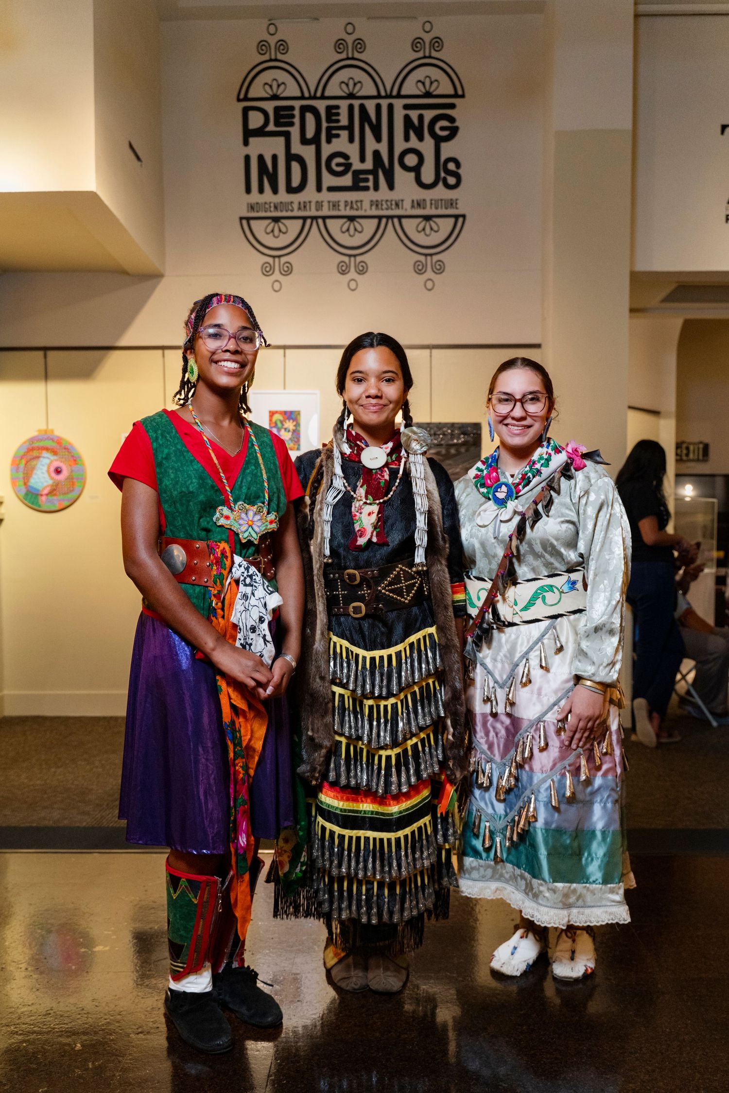 Three people wearing traditional regalia and colorful clothing pose together at an Indigenous arts event.