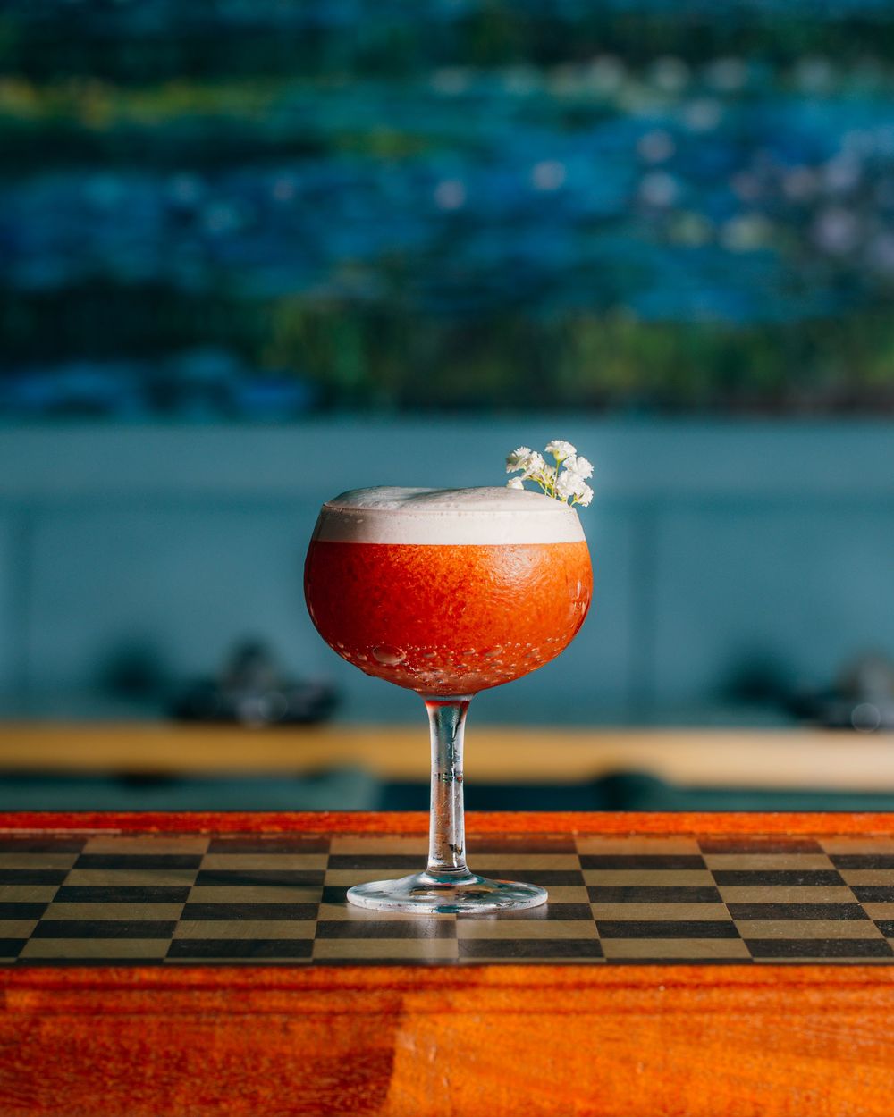 A vibrant red cocktail garnished with small white flowers sits in a coupe glass on a wooden checkered surface.