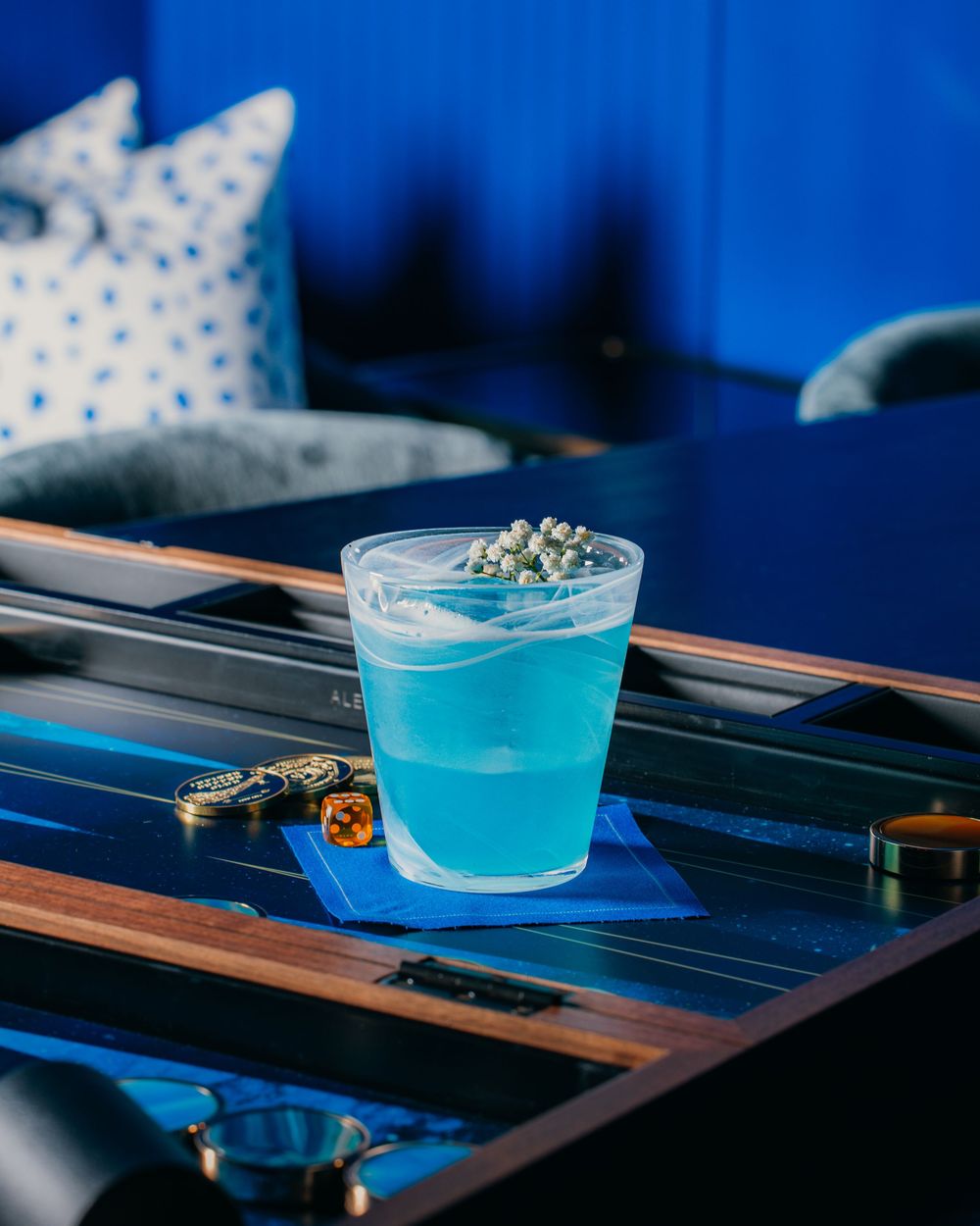 A vibrant blue cocktail in a small glass cup sits on a dark wooden surface with dramatic lighting.