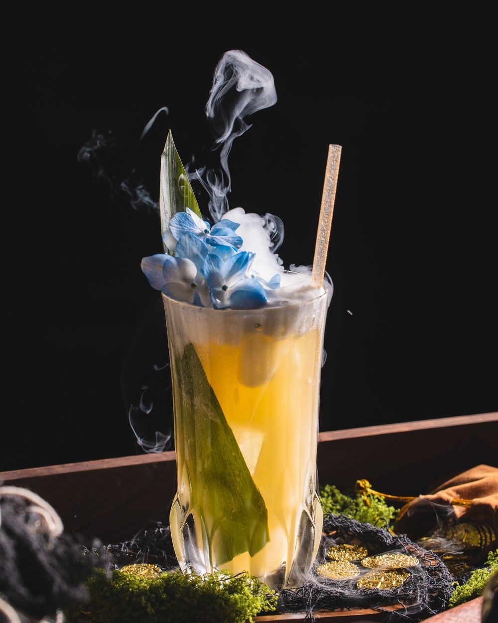 A dramatic cocktail with yellow liquid and blue smoke effect garnished with herbs on a dark background.