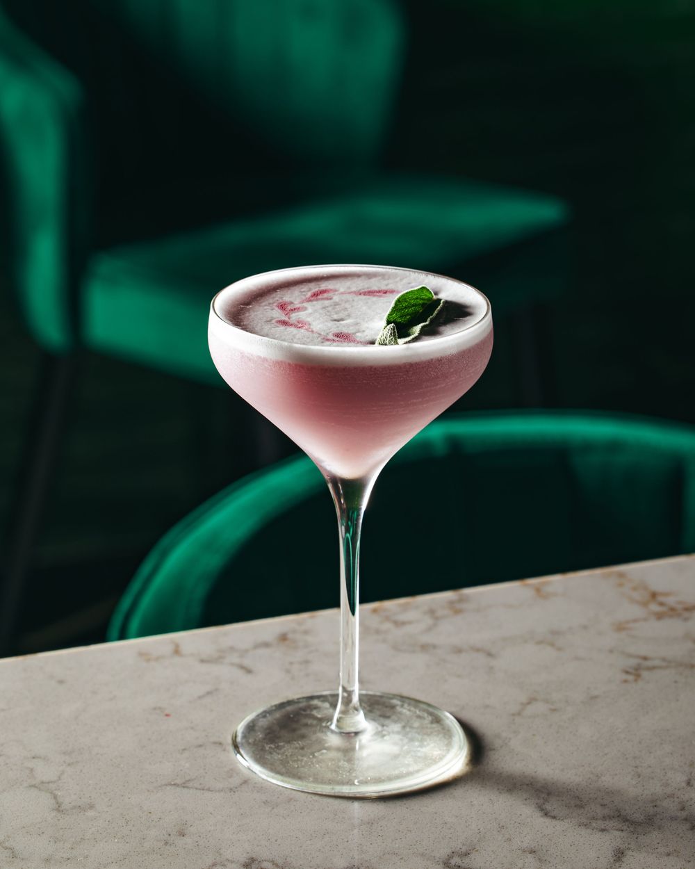 Pink cocktail in coupe glass garnished with mint leaf against dark green background.