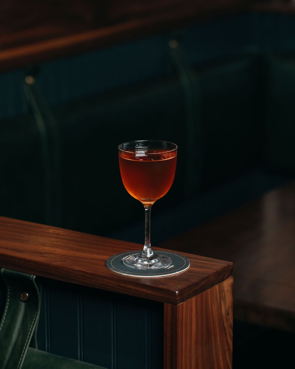 Dark amber cocktail in a coupe glass on wooden surface with moody lighting.