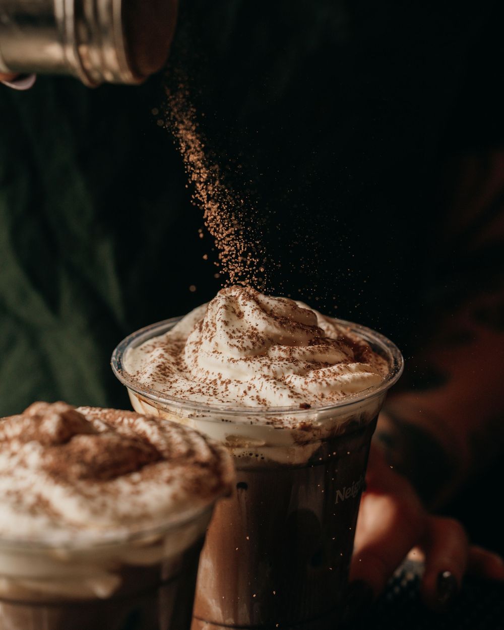 A sequence of whipped cream desserts being dusted with cocoa powder in glass cups against a dark background.