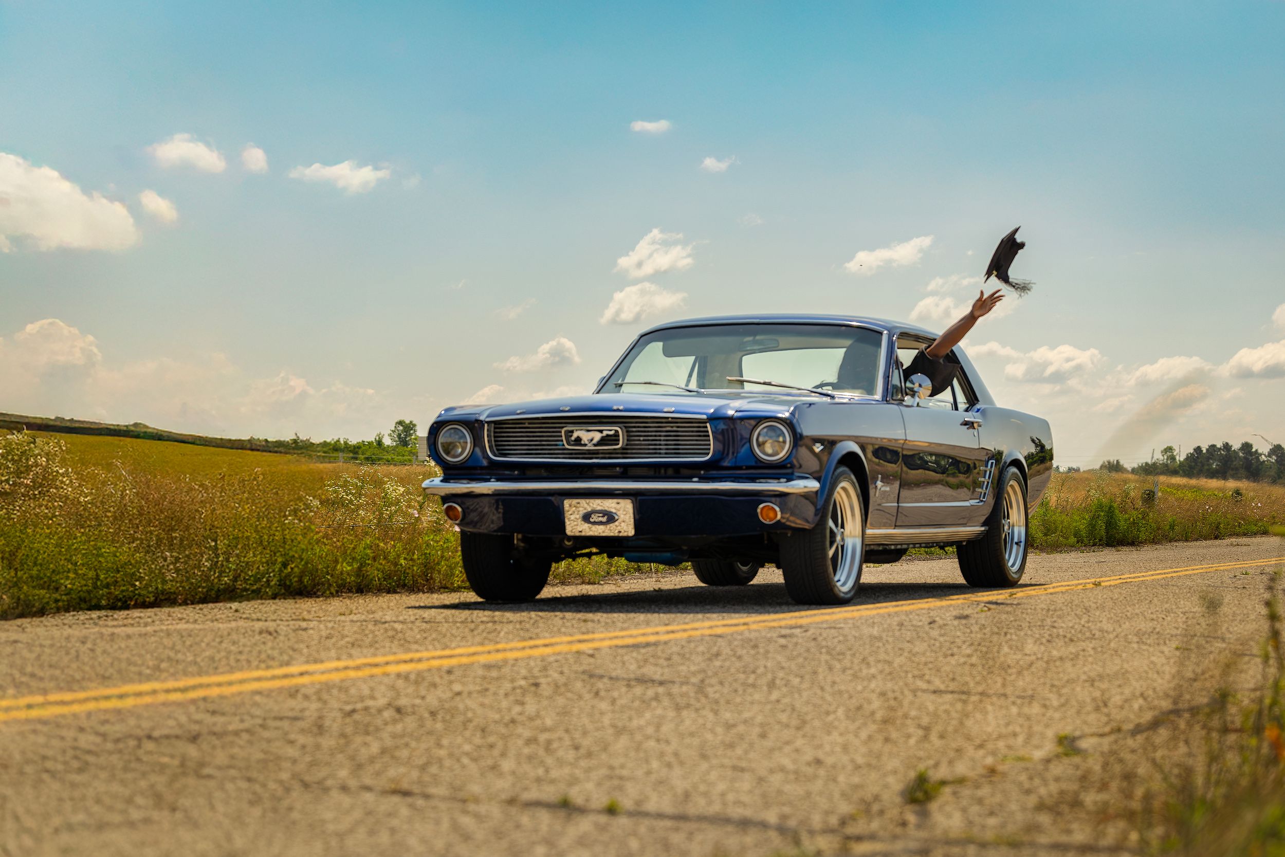 A classic dark blue Ford Mustang driving down a rural highway against a blue sky with puffy clouds.